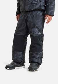 Schwarze Outdoor-Hose mit Muster, die eine Mischung aus strukturierten und glatten Materialien aufweist, versehen mit Reißverschlusstaschen und elastischen Bündchen.