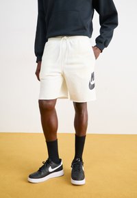 Vita träningsshorts med elastisk midja och snöre, med en svart Nike-logotyp på vänster sida, tillsammans med svarta sneakers och strumpor.