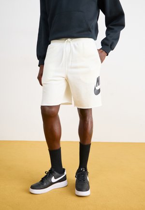 Vita träningsshorts med elastisk midja och snöre, med en svart Nike-logotyp på vänster sida, tillsammans med svarta sneakers och strumpor.