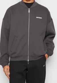 Graue Bomberjacke mit einem Reißverschluss an der Vorderseite, geripptem Kragen und Bündchen. Mit einem weißen Logo auf der linken Brust und seitlichen Taschen.