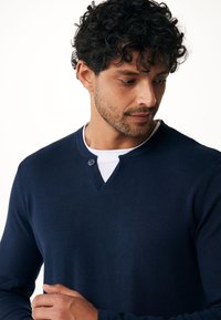 Top a maniche lunghe blu navy con scollatura a V e dettaglio a bottoni. Indossato sopra una t-shirt bianca a girocollo, che mette in evidenza una morbida trama a maglia.
