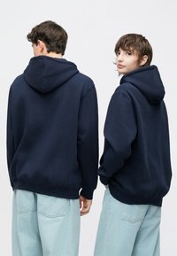 Tamsiai mėlynas gobtuvas su laisvu siluetu, ribotais rankogaliais ir apačia. Dėvimas ant šviesiai mėlyno džinso, pasižymi lygiu paviršiumi ir minimalistiniu dizainu.