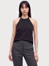 Junge Frau mit glatten dunklen Haaren, die ein ärmelloses schwarzes Neckholder-Top und hoch taillierte graue Hosen trägt, steht vor einem schlichten hellen Hintergrund.