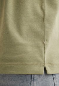 Gros plan de l'ourlet d'une chemise vert olive clair avec une fente latérale et un petit détail "V" cousu, portée sur un jean en denim bleu clair.