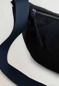 Czarna torba noszona na przekroju z teksturowanym granatowym paskiem i srebrnym detalem w postaci suwaka. Charakteryzuje się zaokrąglonym kształtem i krótkim suwakiem.