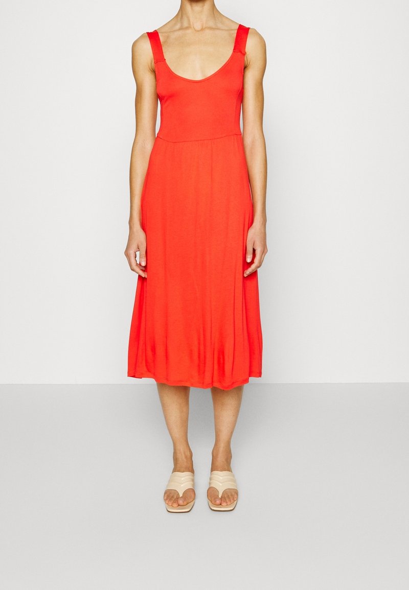 Robe rouge sans manches avec un col arrondi, un corsage ajusté et une jupe évasée. Le tissu semble lisse et doux, s'harmonisant avec des sandales beiges à enfiler.