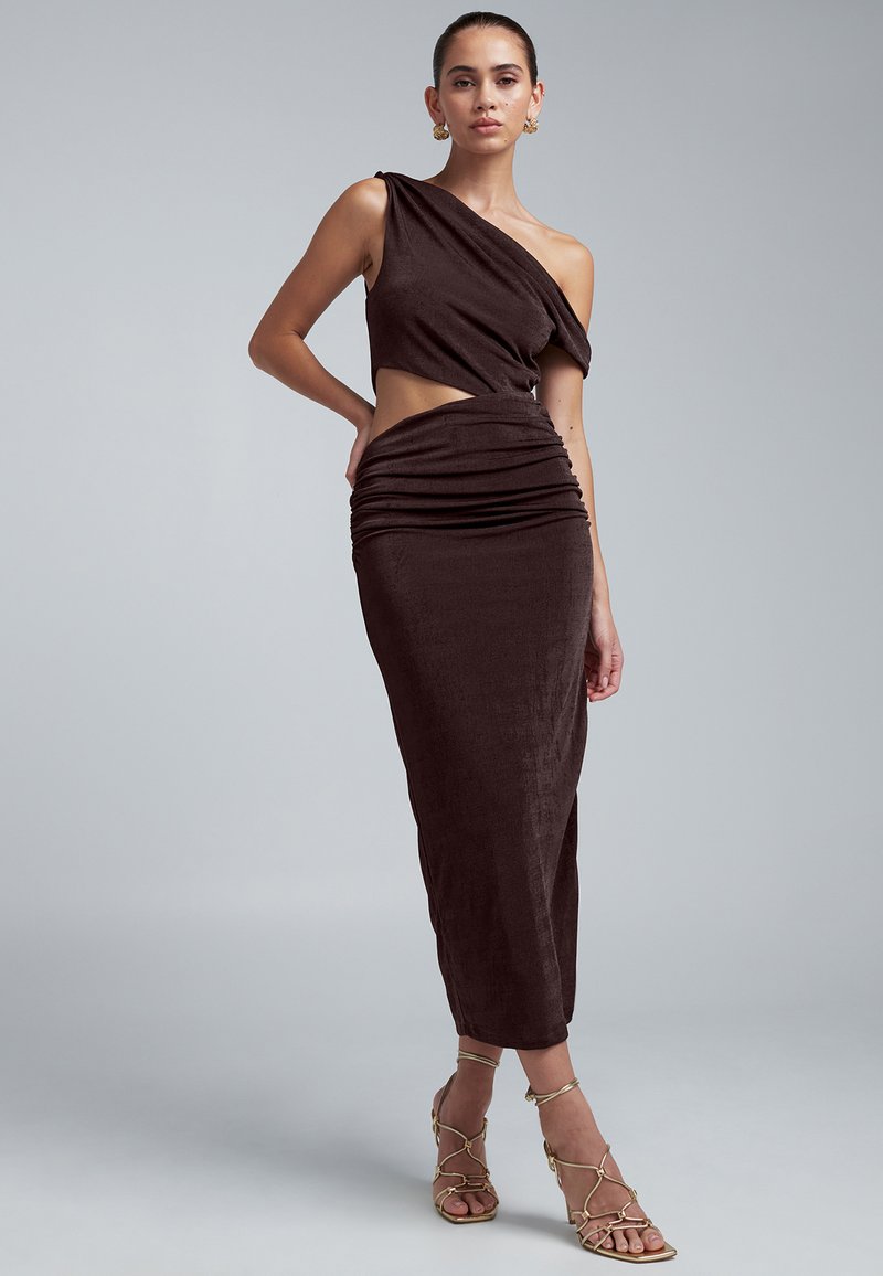 Donna che indossa un abito marrone scuro monospalla con ritagli e vita arricciata, abbinato a sandali con cinturini dorati e tacco, e orecchini dorati, in piedi davanti a uno sfondo grigio.