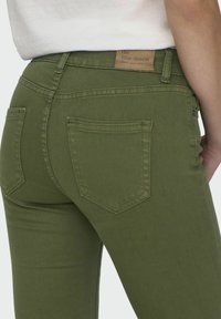 Jean en denim vert avec un design classique à cinq poches, des passants de ceinture et une étiquette en cuir sur le dos de la taille avec les détails de la marque.