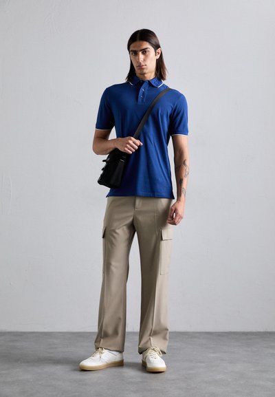 Bărbat cu păr până la umeri, îmbrăcat cu un tricou polo albastru, pantaloni cargo bej, adidași albi și o geantă de umăr neagră, stând lângă un perete simplu.