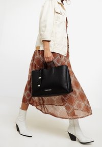 Czarna skórzana torebka na podwójnych uchwytach, gładka faktura, srebrne akcenty i drukowane logo. Połączona z wzorzystą sukienką i białymi botkami za kostkę.