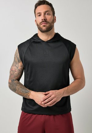 Uomo con barba e braccio sinistro tatuato che indossa una canotta nera con cappuccio e pantaloncini rossi, in piedi con le mani intrecciate.