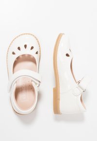 Białe lakierowane buty typu Mary Jane z okrągłym noskiem, wyciętym kwiatowym wzorem, paseczkiem wokół kostki oraz beżową gumową podeszwą. Gładka tekstura.