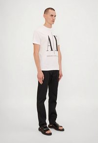 Jovem de pé, vestindo uma t-shirt branca da Armani Exchange, calças pretas e sandálias pretas de dedo frente contra um fundo branco liso.