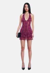 Vinröd halterneckklänning med spetsöverlägg, djup urringning, transparenta partier och volangkant. Modellen håller en liten svart kuvertväska och bär svarta klackar.