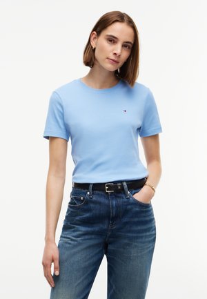 Jeune femme en chemise à manches courtes bleu clair avec un petit logo, jean bleu, ceinture noire et bracelet en or, debout avec une main dans la poche.