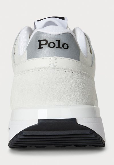 Apskatot no aizmugures baltās zamšādas un auduma sporta kurpes ar melno "Polo" logo uz sudraba papēža un melno gumijas zoli.