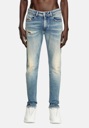 Männliches Model trägt ausgewaschene blaue, abgenutzte Skinny-Jeans mit leichten Rissen am Oberschenkel und Knie, kombiniert mit weißen Sneakers.