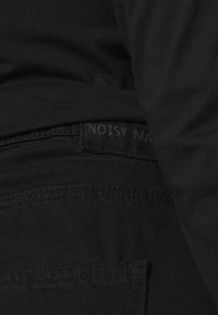 Jeans in denim nero con un design elegante. Presentano un'etichetta visibile con la scritta "NOISY MAY" e dettagli di cucitura sottili. Texture liscia.