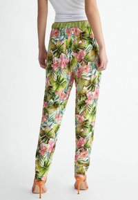 Woman wearing white top and tropical floral print pants with green waistband, paired with orange high-heeled sandals, standing facing back.