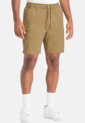 Pantalones cortos caqui de tela tejida, con cinturilla elástica y cordón ajustable, bolsillos laterales y un corte relajado. Llevados con zapatillas blancas.