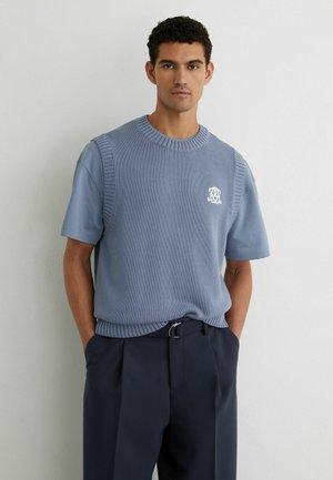 Mann trägt hellblauen gestrickten ärmellosen Pullover über einem passenden kurzärmligen Hemd mit dunkler plissierter Hose, steht vor weißer Wand.