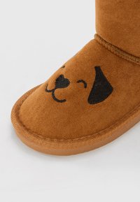 Bruna mockasiner i mocka med rund tå, prydda med en svart broderad hundansiktsdesign och svarta öronaccenter. Mjuk textur och sydda kanter.