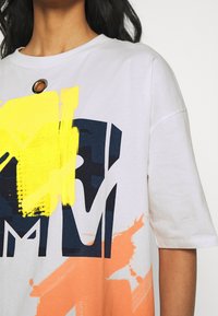 Bílé oversized tričko s abstraktními žlutými, námořnicky modrými a oranžovými grafikami. Má kulatý kovový akcent u límce.