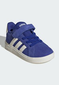 Modré dětské tenisky Adidas s bílými pruhy, suchým zipem, tkaničkami a texturovanou bílou podrážkou na světlém pozadí.