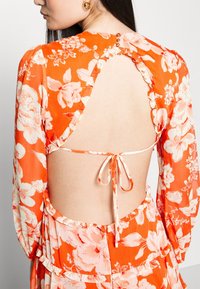 Robe florale orange avec une découpe profonde dans le dos, fermeture à nouer et bords volantés. Présente de grandes fleurs blanches et des accents de boutons au niveau du cou.
