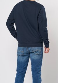 Sweat-shirt marine à manches longues, poignets et ourlet côtelés ; associé à un jean bleu délavé avec poches arrière et légère usure.