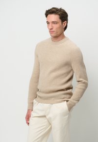 Beige ribbad stickad tröja med rund halsringning och åtsittande ärmar, bärs med ljusa byxor. Mjuk textur, enkel design.