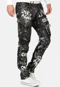 Pantalons cargo noirs avec éclaboussures de peinture blanche, texte graphique et accents cloutés. Dispose de poches et de sangles ajustables pour un design unique.