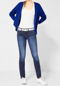 Blå öppen cardigan över en vit linne, ihopparat med mörkblå slim-fit jeans och vita sneakers, med ett ljust färgad bälte.