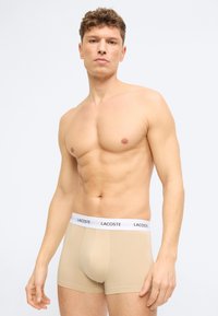 Άνδρας χωρίς πουκάμισο με κοντά σγουρά μαλλιά, ντυμένος με μπεζ μπόξερ της Lacoste με λευκή ζώνη μέσης με λογότυπο, στέκεται μπροστά σε ένα απλό φωτεινό φόντο.