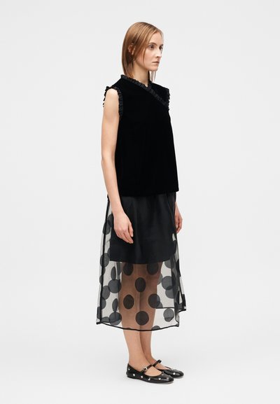 Svart sammetslinne utan ärmar med rynkade kanter, kombinerat med en genomskinlig svart kjol prydd med stora cirkulära prickar.