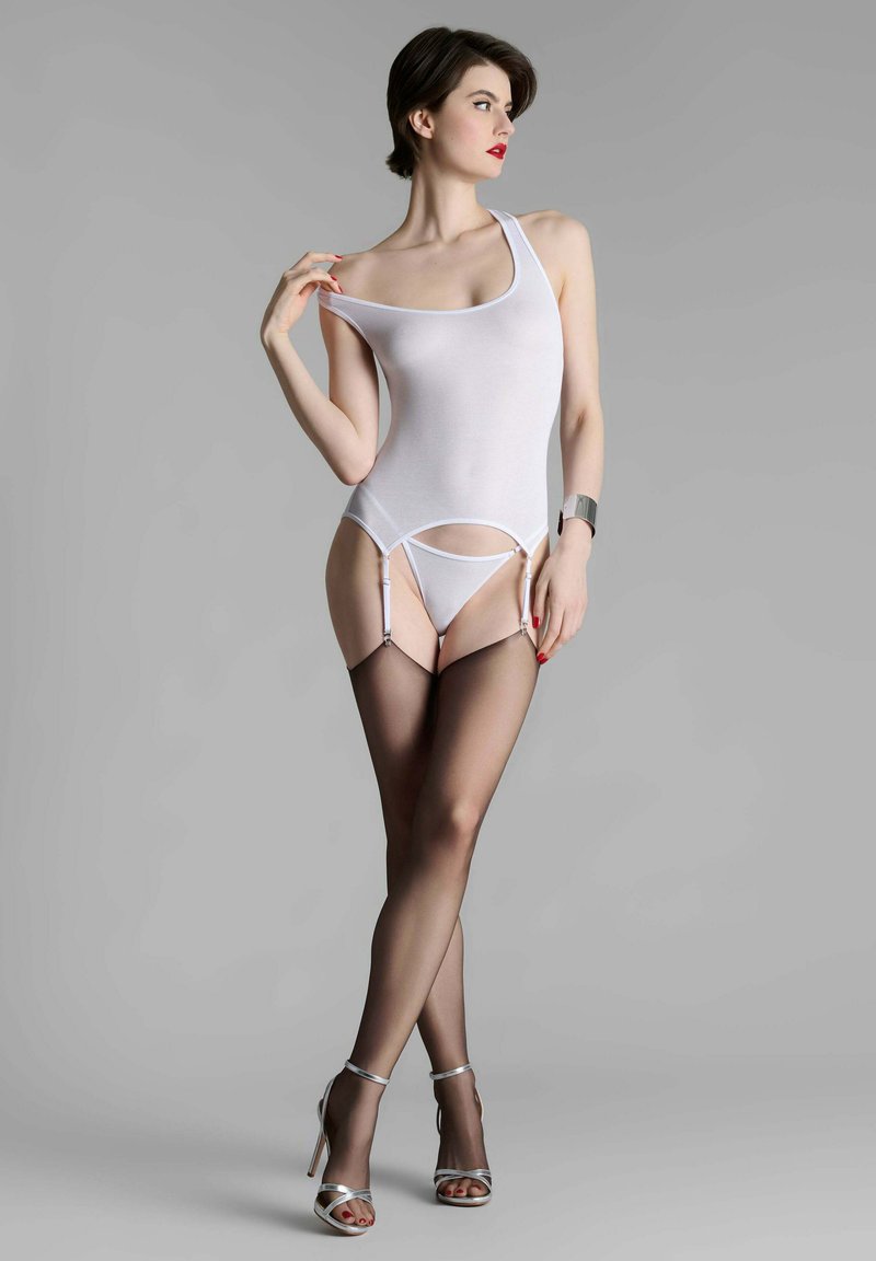 Femme posant dans un justaucorps blanc avec jarretelles, bas noirs transparents, talons aiguilles argentés, rouge à lèvres rouge et cheveux courts foncés sur un fond gris.