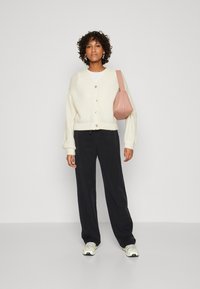 White knitted cardigan with buttons, black loose-fitting pants, and a pink shoulder bag. Footwear features a white and yellow design.