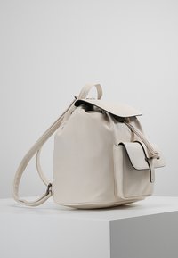 Biały skórzany plecak o zaokrąglonym kształcie, z zamknięciem na sznurek oraz przednią kieszenią z klapą i kontrastowym wykończeniem. Gładka tekstura.