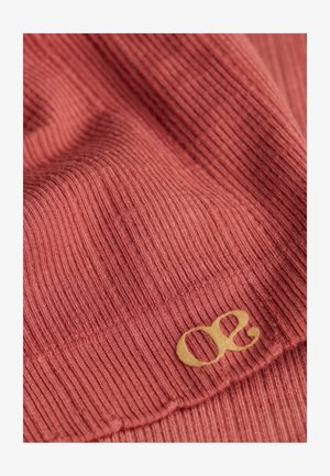 Tissu côtelé dans une couleur rougeâtre atténuée avec un accent de logo doré. Texture lisse avec un détail de bord ondulé sur le tissu.