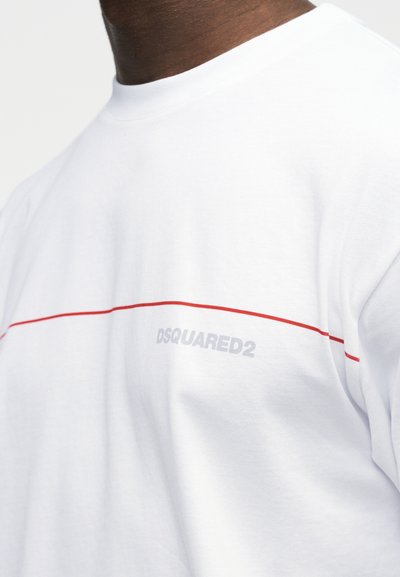 Κοντινό πλάνο ενός ατόμου που φοράει λευκό μπλουζάκι με λεπτή κόκκινη οριζόντια γραμμή και την επιγραφή "DSQUARED2" σε ανοιχτό γκρι στο στήθος.