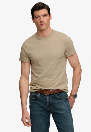 Mann trägt beige T-Shirt mit kurzen Ärmeln, blaue Jeans, braunen Ledergürtel und Armbanduhr, steht mit einer Hand in der Tasche vor weißem Hintergrund.