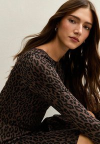 Vestido de estampado de leopardo en marrón y negro, diseño ajustado con mangas largas, tela suave y escote redondo. Modelo posando contra un fondo neutro.