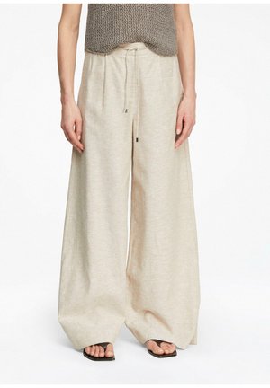 Kobieta w beżowych szerokich spodniach ze sznurkiem, brązowym dzianinowym topie i czarnych sandałach z odkrytymi palcami, stojąca na białym tle.