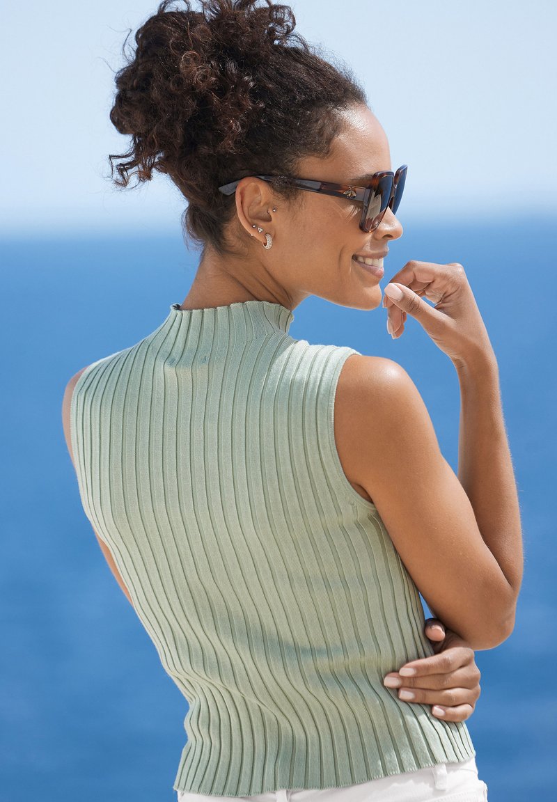 Mintgrünes, geripptes, ärmelloses Oberteil mit hohem Halsausschnitt, das eine figurbetonte Silhouette und eine strukturierte Oberfläche aufweist. Klarer blauer Hintergrund.