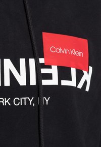 Sudadera negra con texto grande en blanco, una etiqueta rectangular roja con "Calvin Klein" y un cordón texturizado.