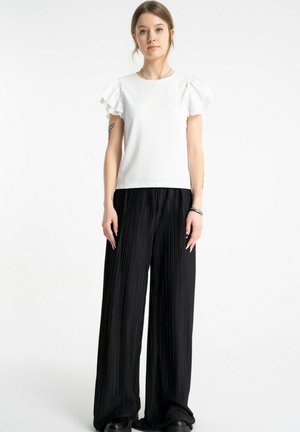 Młoda kobieta o jasnobrązowych włosach ubrana w biały top z falbanami na rękawach i czarne szerokie plisowane spodnie, stojąca na białym tle.