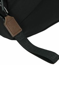 Borsa in tessuto nero con un'etichetta in pelle marrone texture e una tracolla nera intrecciata con lettering in rilievo. La ferramenta è in metallo argentato.