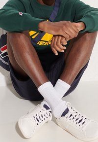 Witte low-top sneakers met ingewikkeld vetersysteem, marineblauwe shorts en een groene trui met een geel logo op de voorkant. Witte kousen zichtbaar.