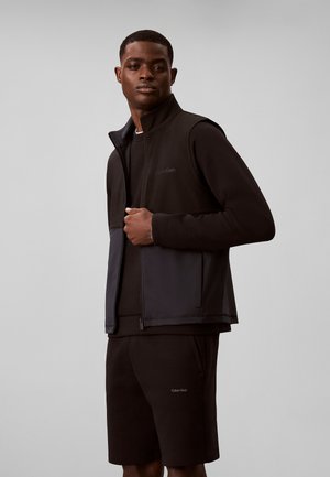 Homme portant un gilet sans manches noir zippé sur une chemise à manches longues et un short noir, debout devant un fond gris uni.