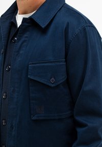 Marineblaue Jacke aus glattem Stoff, mit Knopfverschluss, einer Brusttasche mit Klappe und einem subtilen Logopatch auf der Tasche.
